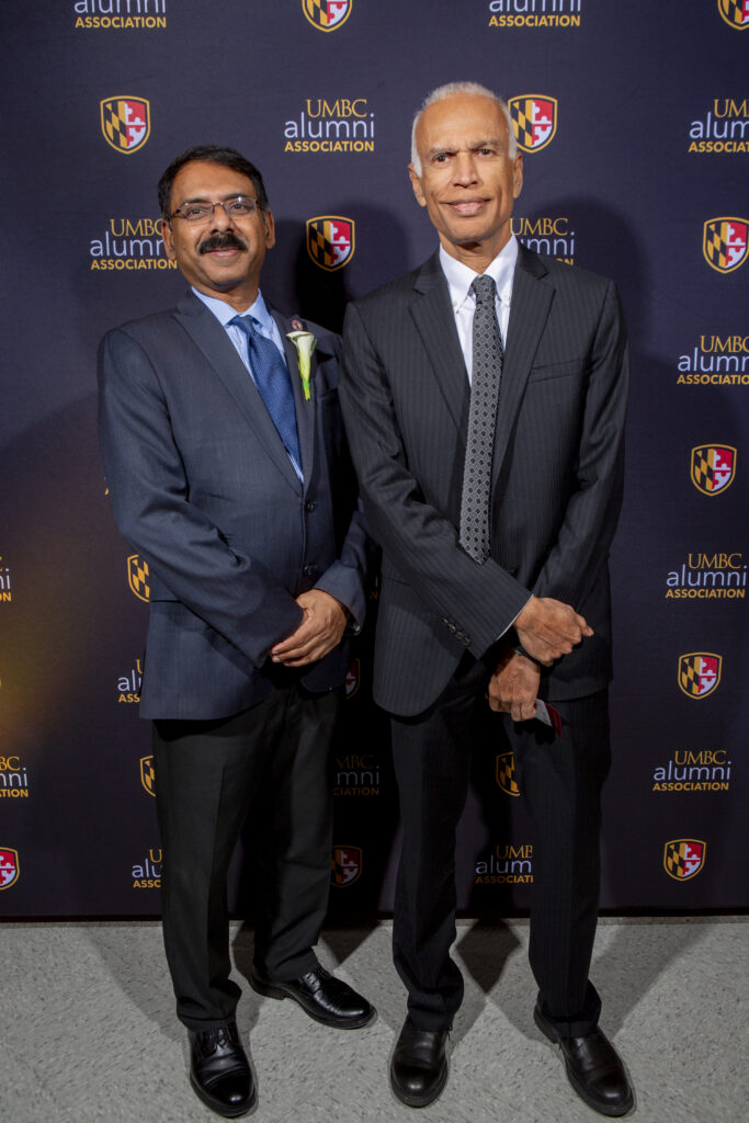 two men in suits, hands clasped in front of them, in front of a banner with the UMBC shield and "UMBC Alumni Association" on it