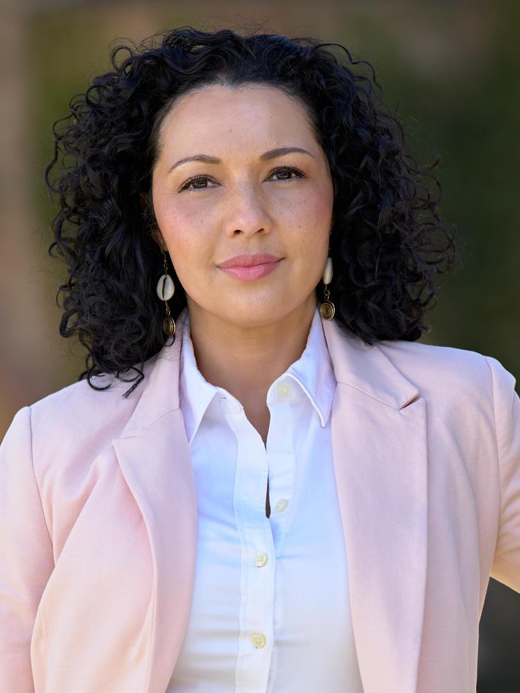 A woman with dark curly hair, wearing a pink jacket and white shirt, smiles at the camera.