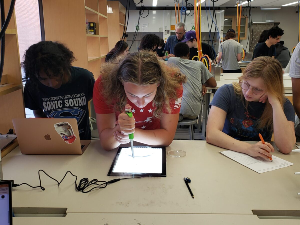 three students lean over a laboratory counter; the one in the center holds a dropper over a backlit pad, one records data on paper in a math class.