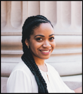 A Black woman will long braided hair, wearing a white, top, smiles at the camera.