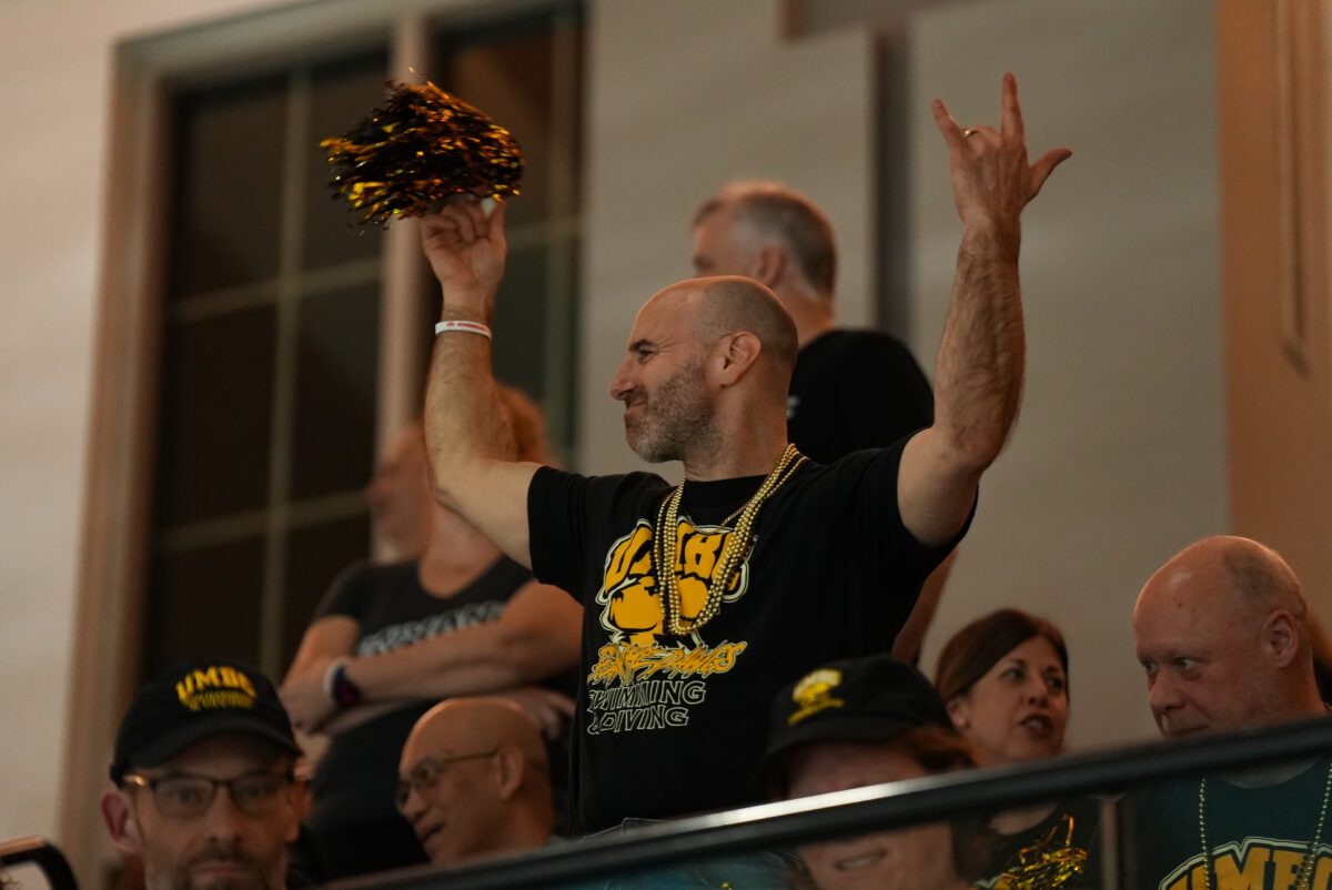 A fan in a UMBC swimming and diving shirt stands with hands raised and a pom-pom in the bleachers 