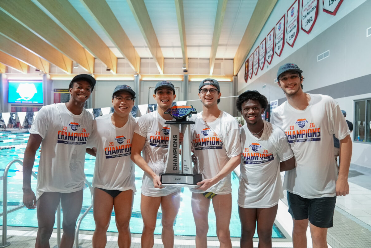 Six men's swimming and diving members from UMBC stand in front of a pool with the America East trophy