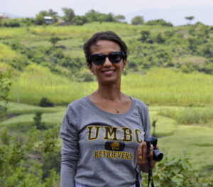 Against a cultivated landscape, a Black woman with short dark hair, wearing a gray shirt that says UMBC, and dark glasses, smiles at the camera.