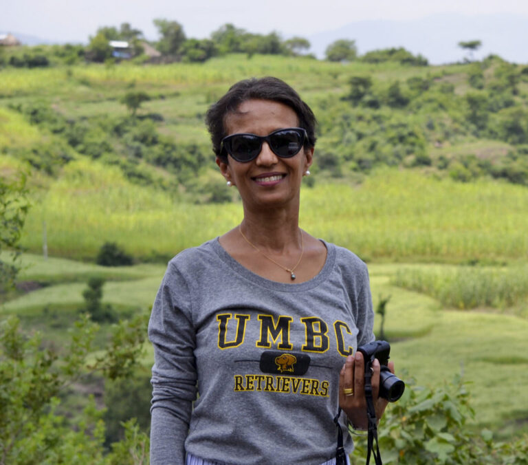 Against a cultivated landscape, a Black woman with short dark hair, wearing a gray shirt that says UMBC, and dark glasses, smiles at the camera.