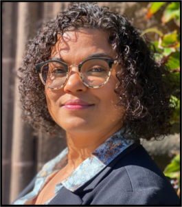 A Black woman with wavy hair and glasses, wearing a blue top and jacket, smiles at the camera.