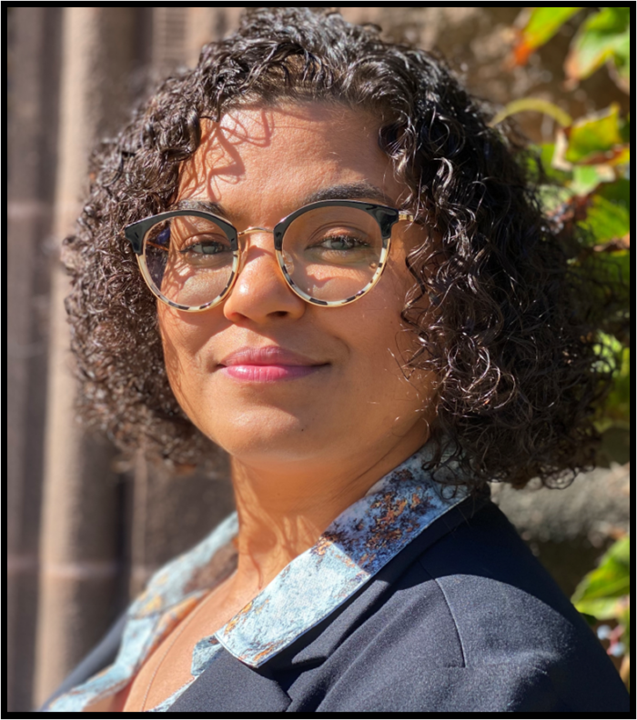 A Black woman with wavy hair and glasses, wearing a blue top and jacket, smiles at the camera.