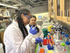 student in lab coat, gloves, and glasses pipets at a lab bench while another student supervises