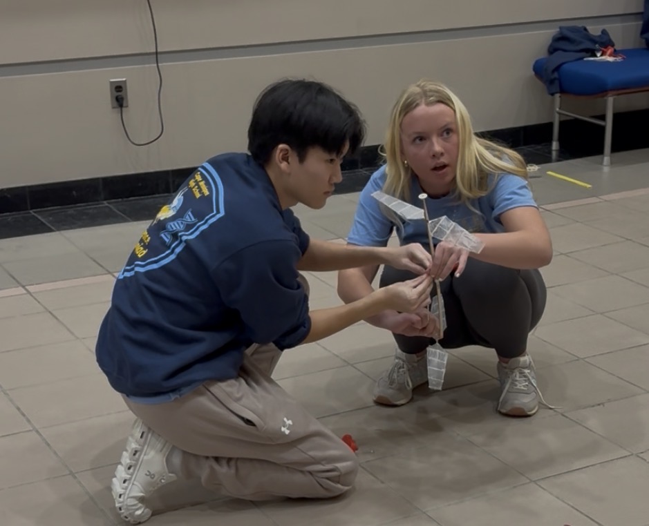 two students kneeling on tile floor, holding a homemade winged device as it prepares to lift off.