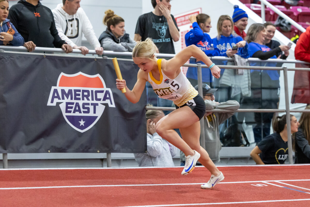 A female student-athlete with blond hair is caught in motion while running an indoor relay race with baton in hand for UMBC