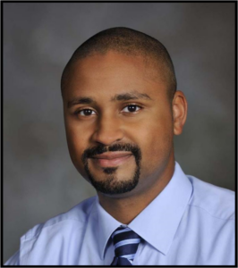 A Black man with a goatee, wearing a blue shirt and tie, smiles at the camera.