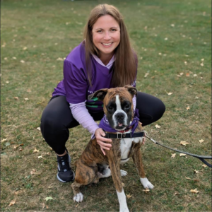woman in purple t-shirt branded with the Lupus Foundation of America, which supports people with the chronic illness, squatting on a grassy field with her hands on her brown and white dog