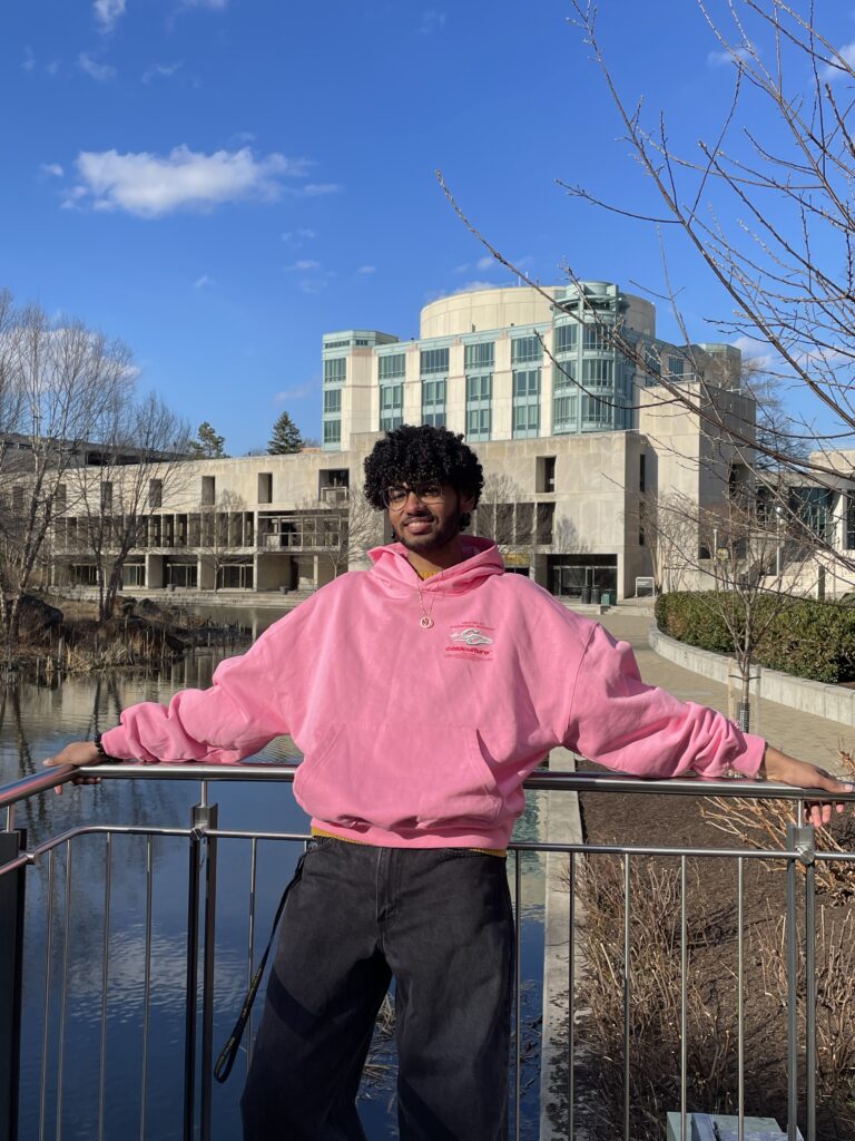 mathematics student leans back on railing in front of pond, large academic building in the background