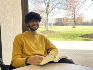 smiling mathematics student in yellow shirt holding a book open and sitting at a table; window behind him shows green grass, trees with spring buds, and a brick academic building