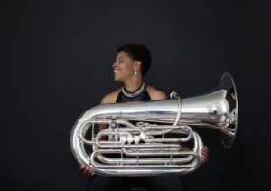 Against a dark background, a Black woman holds a tuba.