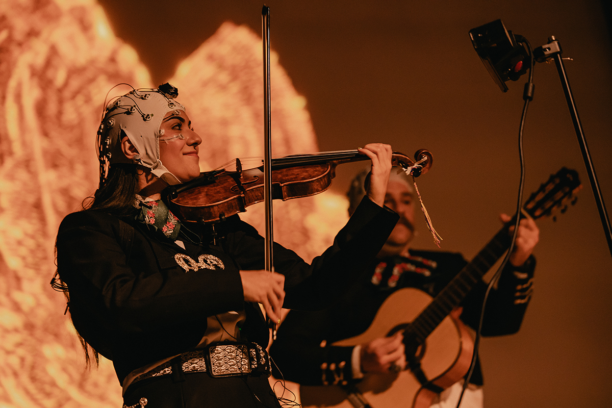 A woman playing violin onstage. She is wearing a headpiece with wires and beside her a man is playing acoustic guitar.