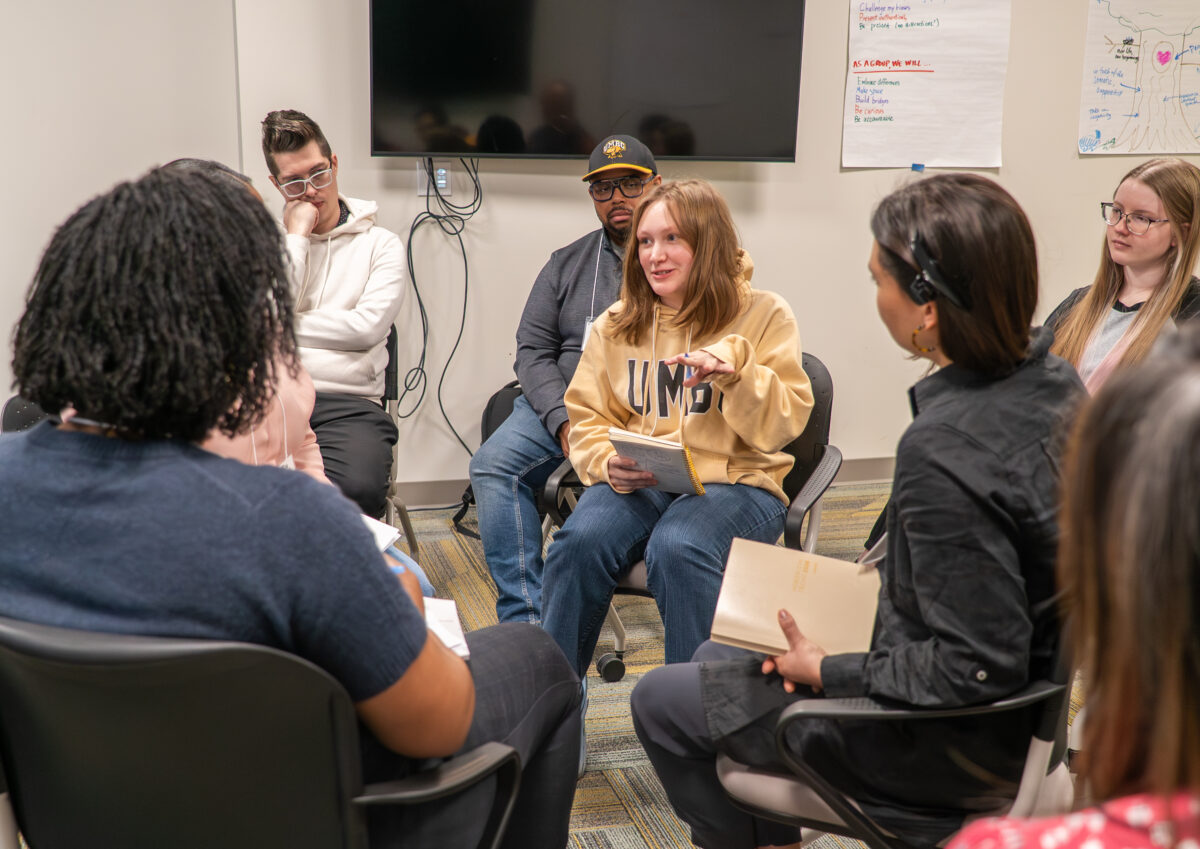 a woman in a yellow UMBC sweatshirt sits in the center of a circle talking about Restorative practices 
