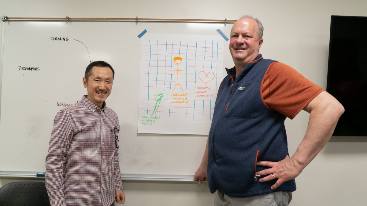 Two men stand it front of whiteboard with a graph taped to it.
