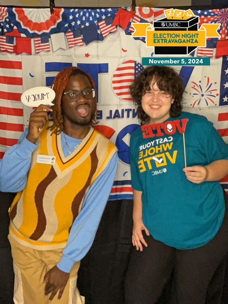 Two people posing in front of a banner, holding up props, urging students to get involved with voting. 