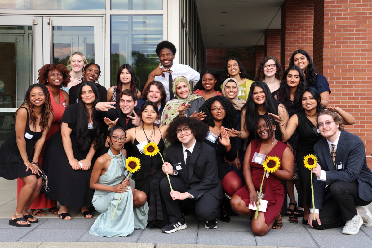 A large group of of students in fancy dress standing outside in front of a building with sunflowers. 
