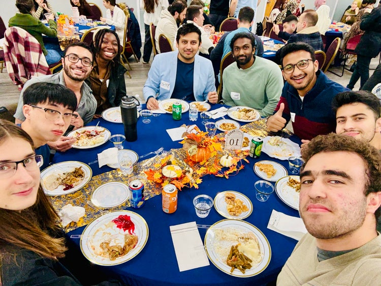 A group of students sit around a table with a blue tablecloth and various food items 