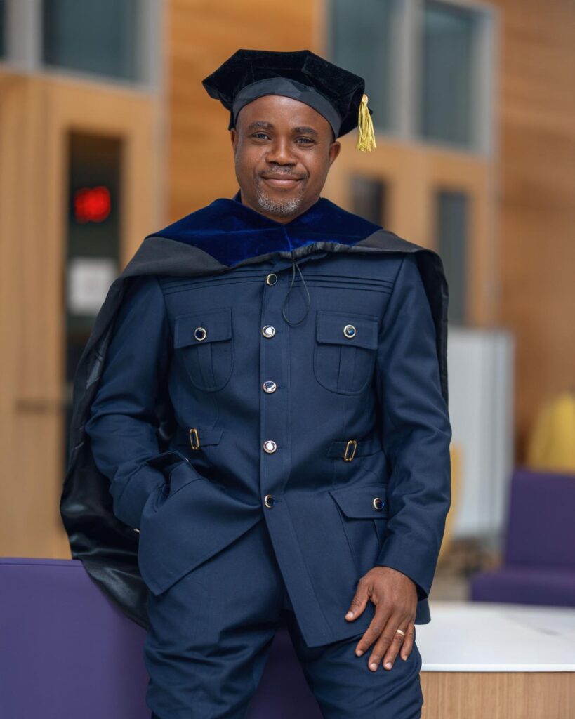 A man stands wearing a dark academic gown with a velvet cap and tassel, posing indoors. He is dressed in a suit beneath the gown. The background features blurred indoor elements, including windows and light wooden walls.