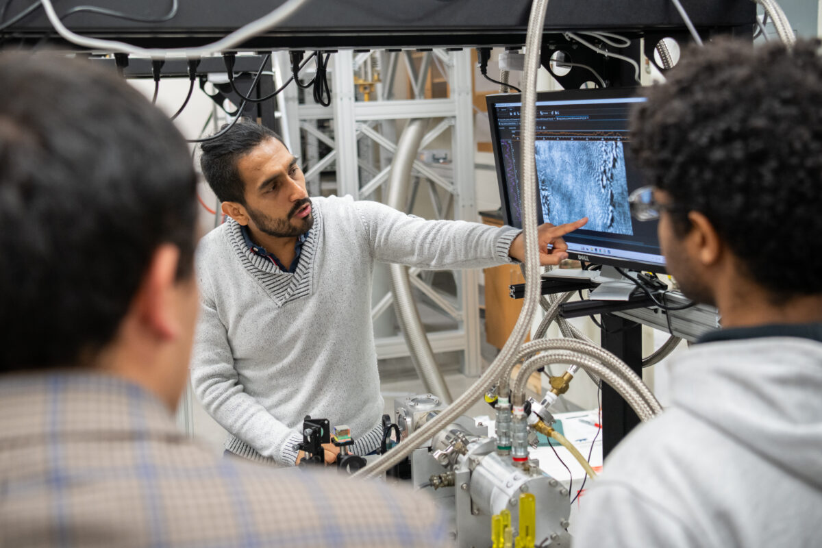 Daniel Suarez-Forero points to a microscope image on a screen while two students look on across a table with an optical laser setup