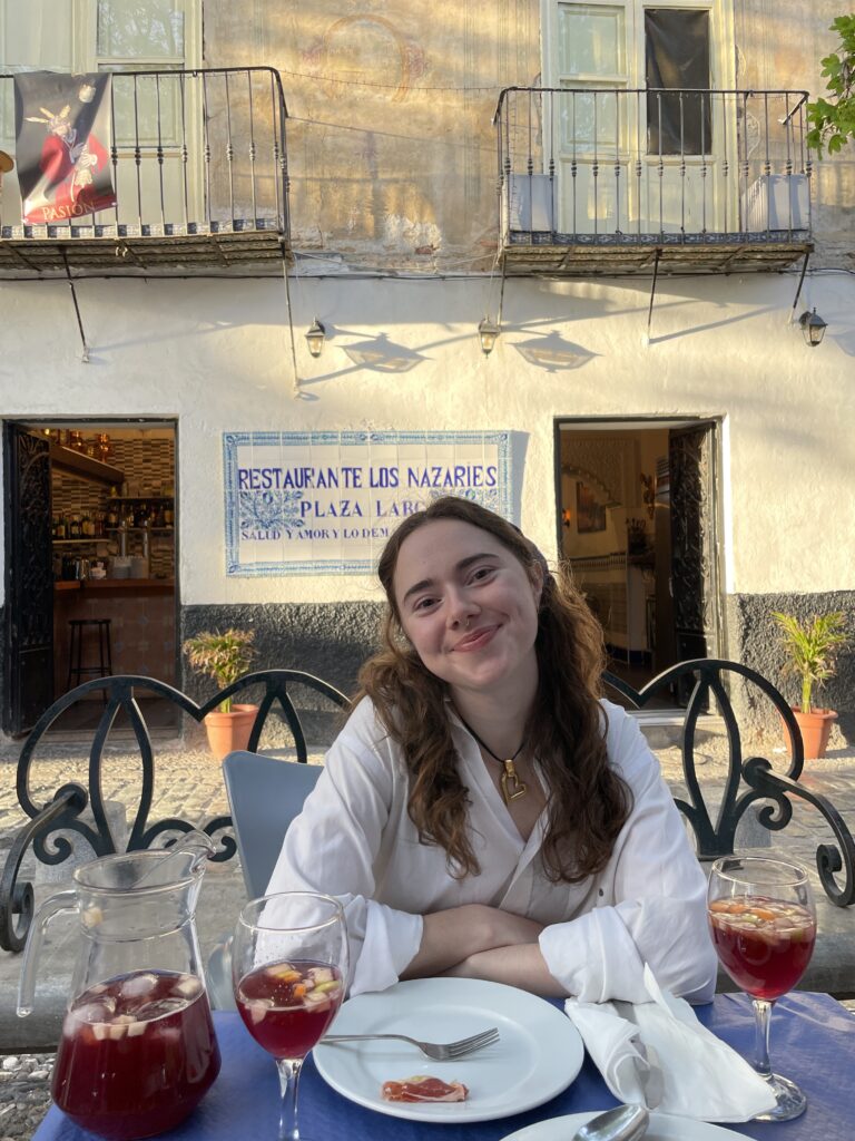 A girl smiles behind a table sitting down with Italian food.