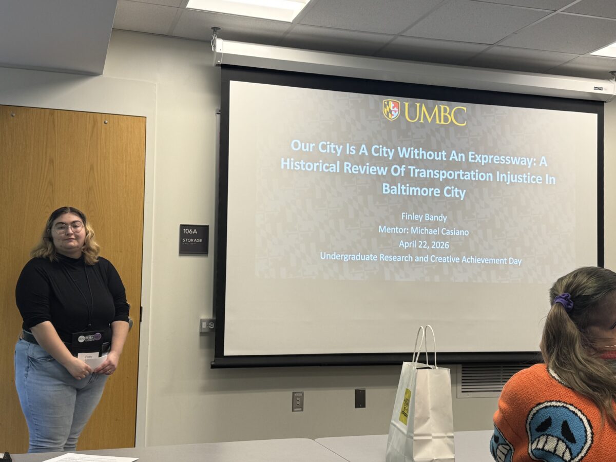 Finley Bandy stands at the front of a college classroom next to a projection screen showing the first slide of their power point presentation on transportation injustice in Baltimore City