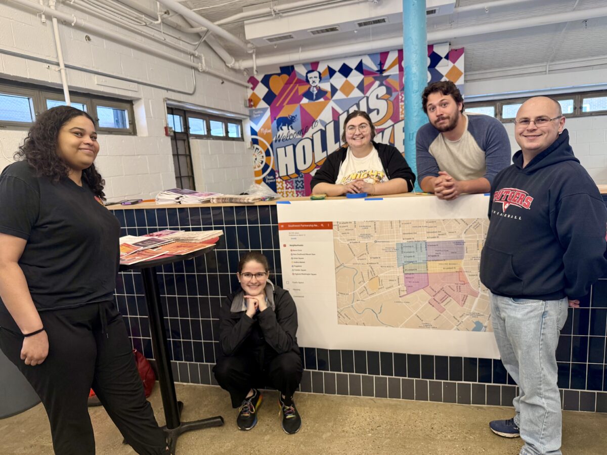 Finley Bandy with five people gathered around a counter with a map of Baltimore hung over the edge and a mural of Baltimore's Hollins Market in the background