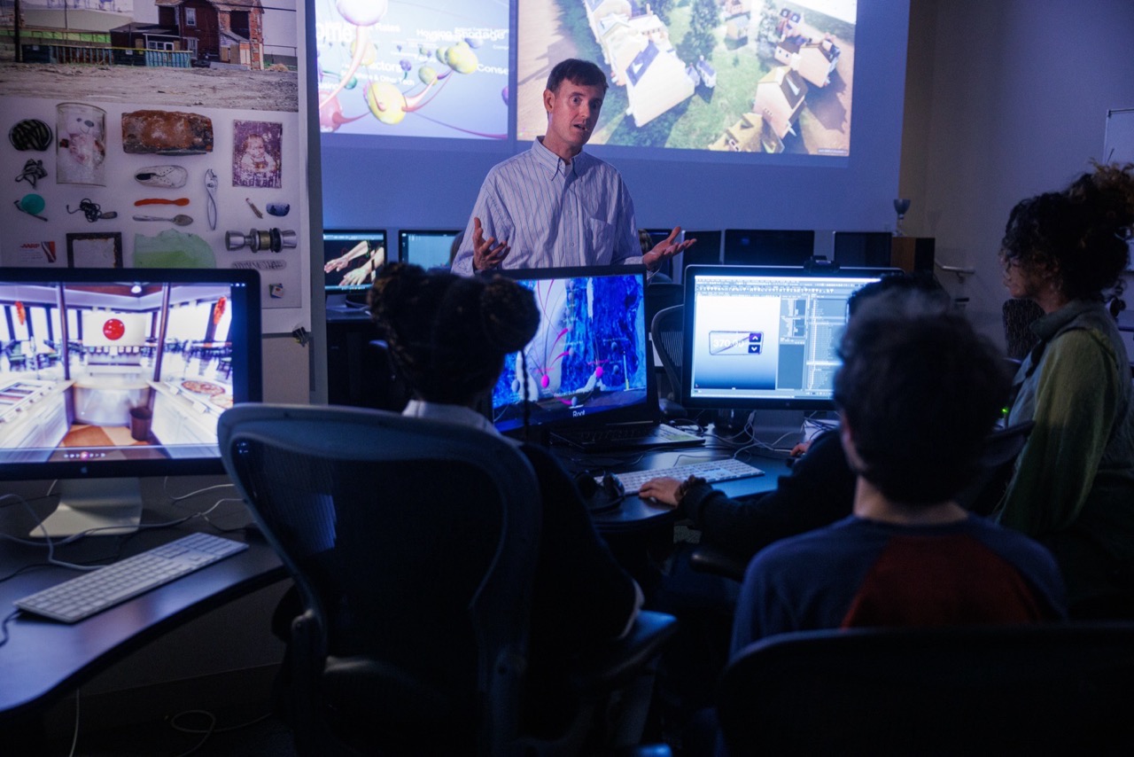 Students work at computers, with an instructor standing near a projection screen.