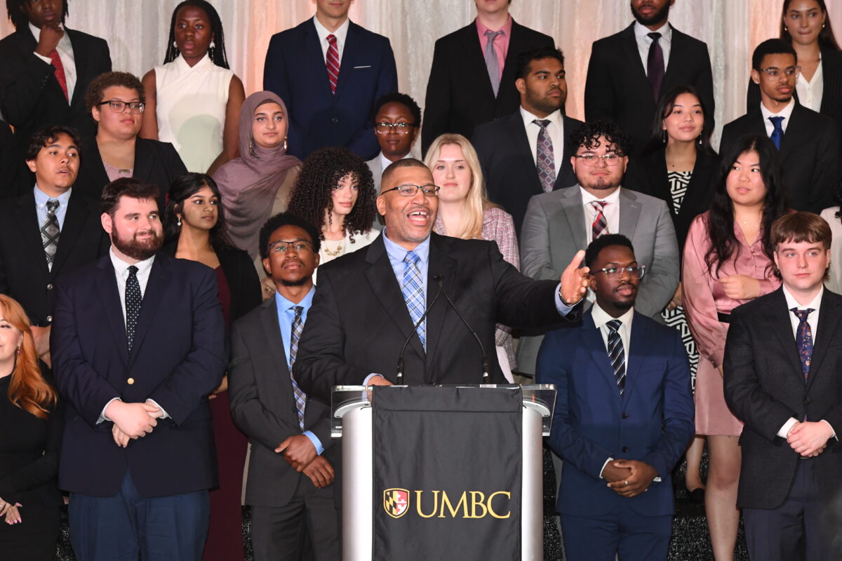 Keith Harmon speaks from a podium, backed by dozens of smartly dressed Meyerhoff Scholars