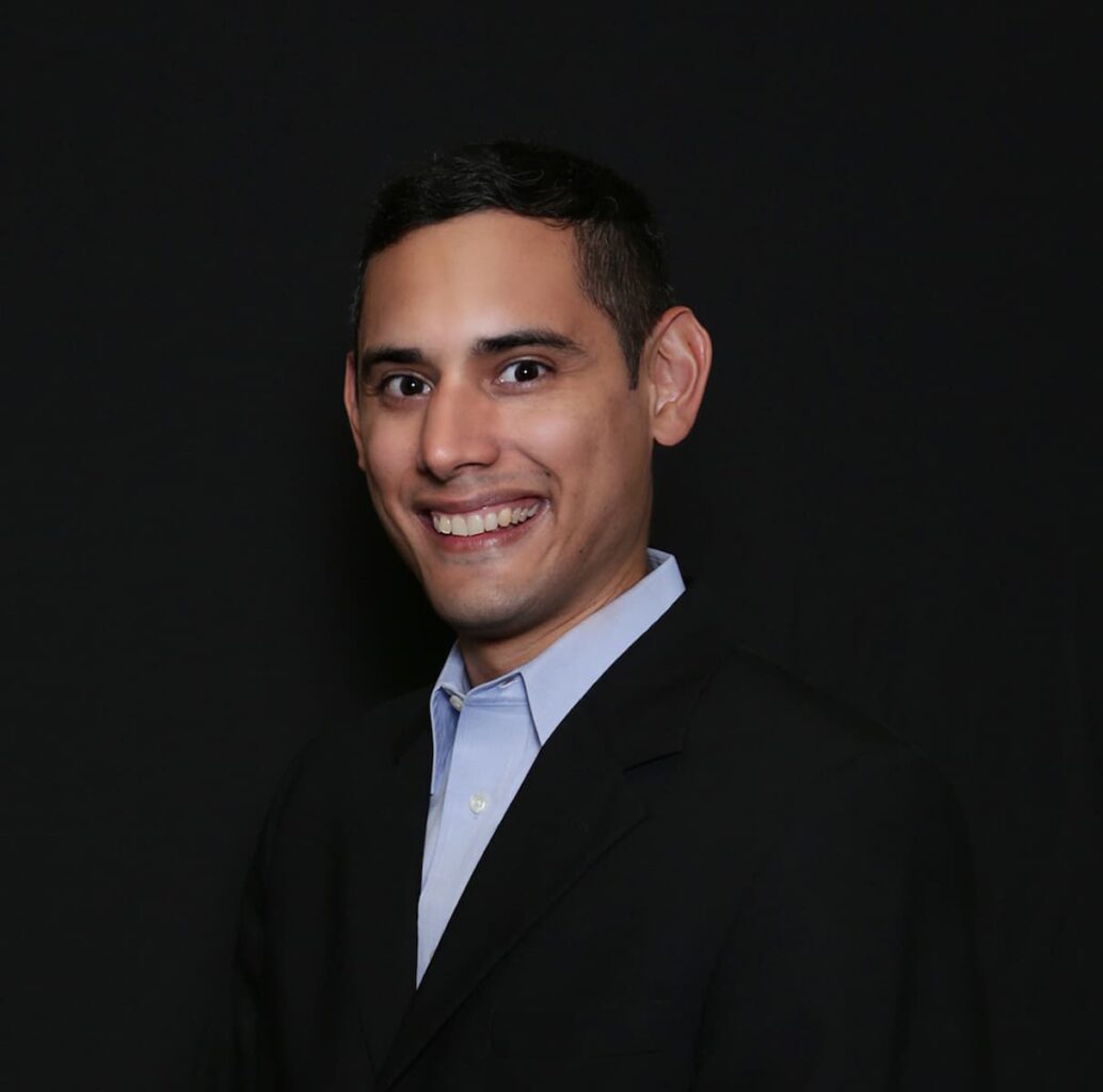 Headshot of a person smiling, wearing a blue button-down shirt and a black suit jacket.