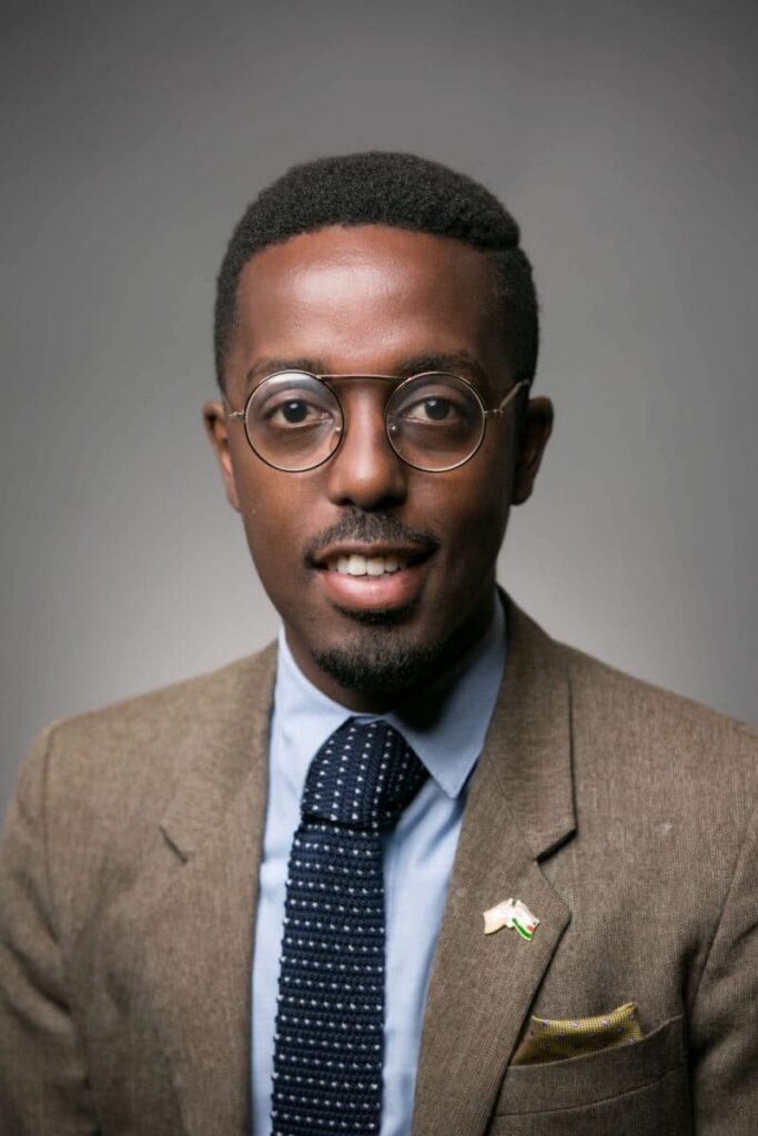 Headshot of a person wearing round glasses, a blazer, and a tie with a small flag pin on the lapel.
