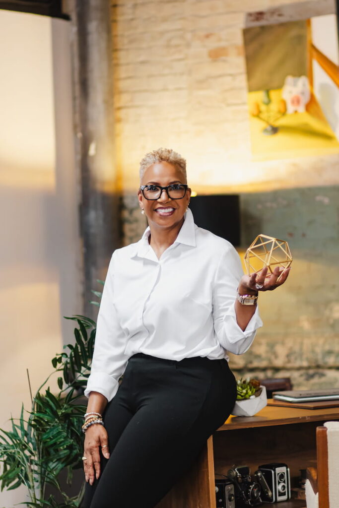 Headshot of a person in glasses holding a gold geometric object. They are wearing a white button-down shirt and sitting on a wooden shelf.