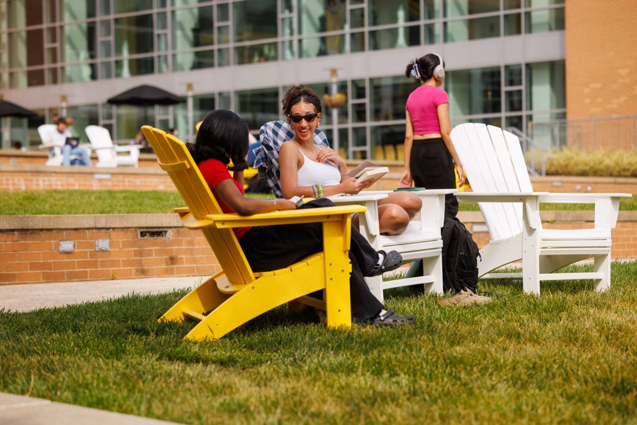 Students relax in Adirondack chairs on the lawn. One student reads a book.