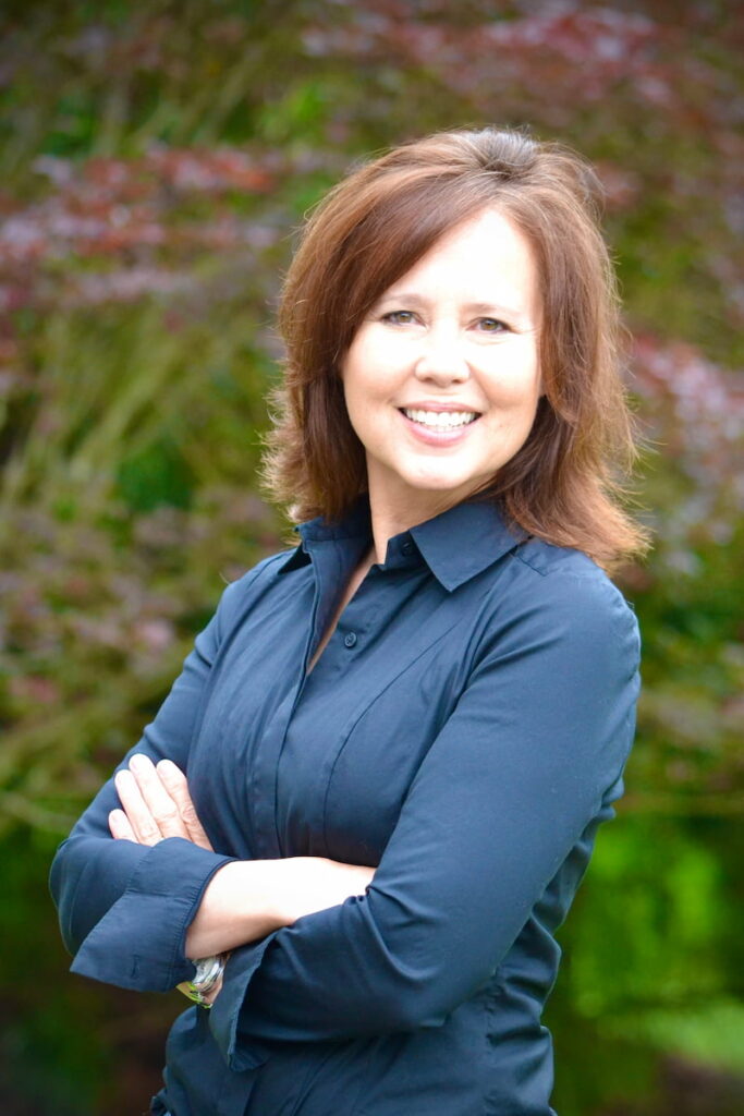 Headshot of a person smiling, with crossed arms. The outdoor setting includes trees and foliage.