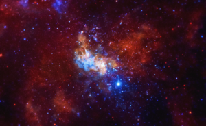 A Chandra X-ray Observatory image around the Sagittarius A* supermassive black hole, located in the center of the Milky Way galaxy, is shown. The image is dominated by vibrant red and blue hues, with a large amount of celestial objects visible. At the center of the image, there is a bright, lumpy object showing the intense X-ray radiation emanating from the Sgr A* black hole. In the surrounding area, there are a couple smaller lumps layered throughout the image, feathering out to a large almost butterfly shape filling much of the screen. The image appears textured, like dozens of blue and orange glow worms are paused in their wriggling. The image offers an unprecedented view of lobes of hot gas extending for a dozen light years on either side of the black hole. These lobes provide evidence for powerful eruptions occurring several times over the last ten thousand years. The image also contains several mysterious X-ray filaments, some of which may be huge magnetic structures interacting with streams of energetic electrons produced by rapidly spinning neutron stars. Such features are known as pulsar wind nebulas.