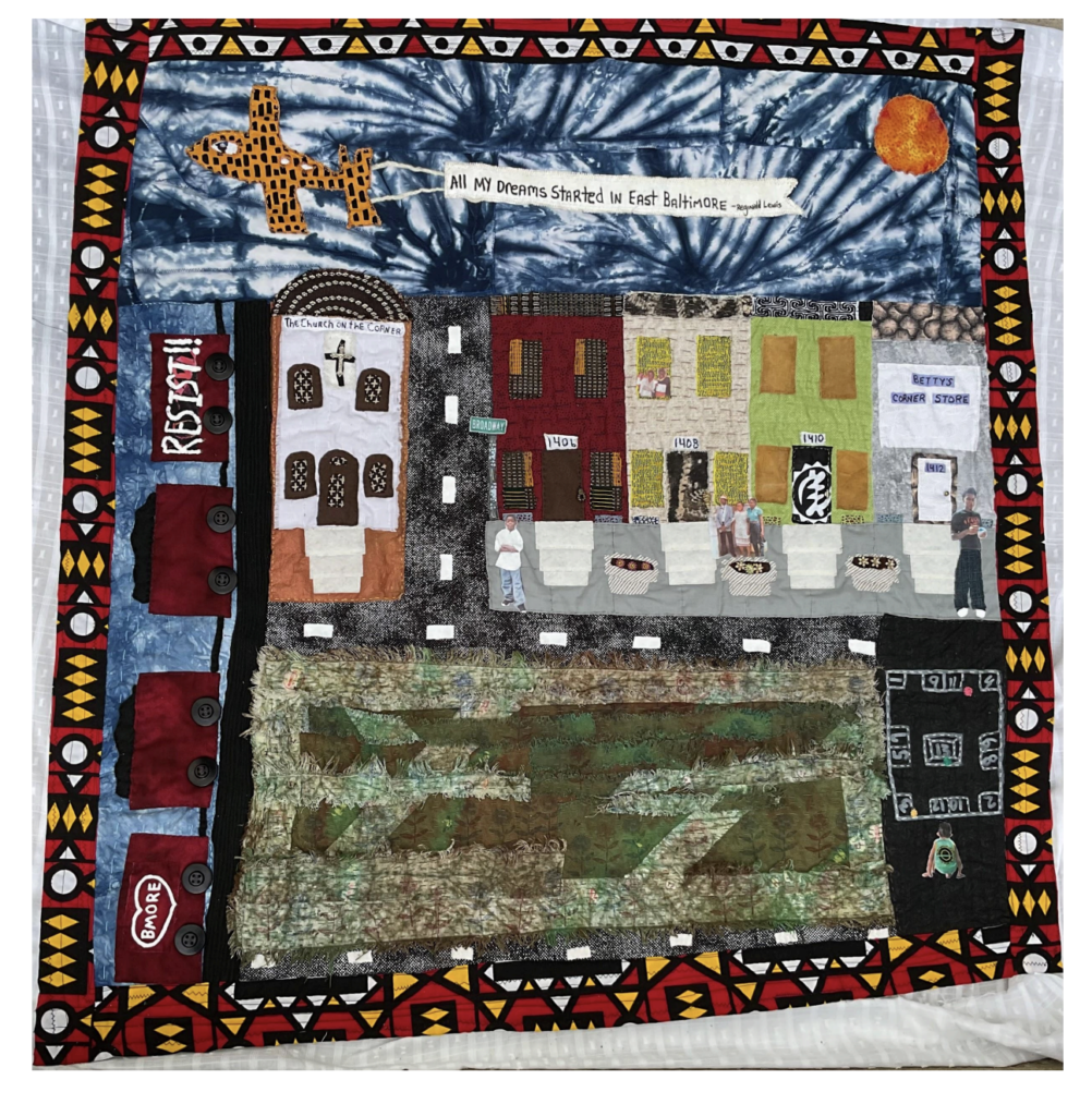 A quilt depicting Baltimore City rowhomes in white, red, green, and beige, with red train cars on the left margin, and a garden in front of the homes. A flying banner has "All My Dreams Started in West Baltimore" stitched at the top of the quilt.