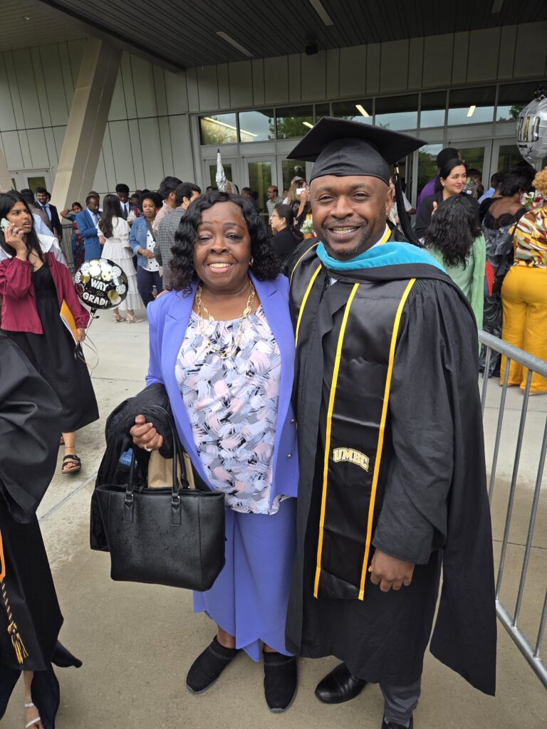 A woman wearing a periwinkle jacket and skirt stands next to her son whose wearing graduation regalia with a crowd of people behind them with graduation balloons