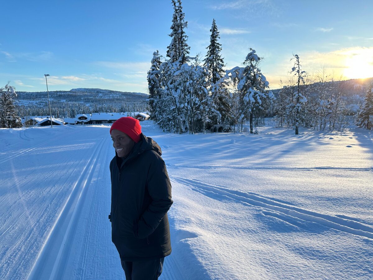 Shanika Freeman, an adult wearing a heavy winter coat and a red stands alone on a snowy field with trees filled with snow and a small village with snow on the roofs behind her