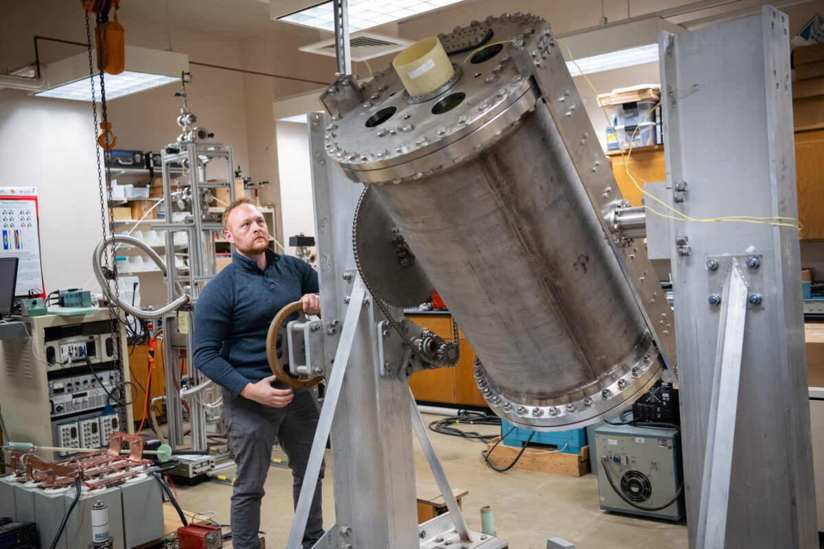 Man stands next to large metallic cylinder mounted on rotatable platform.