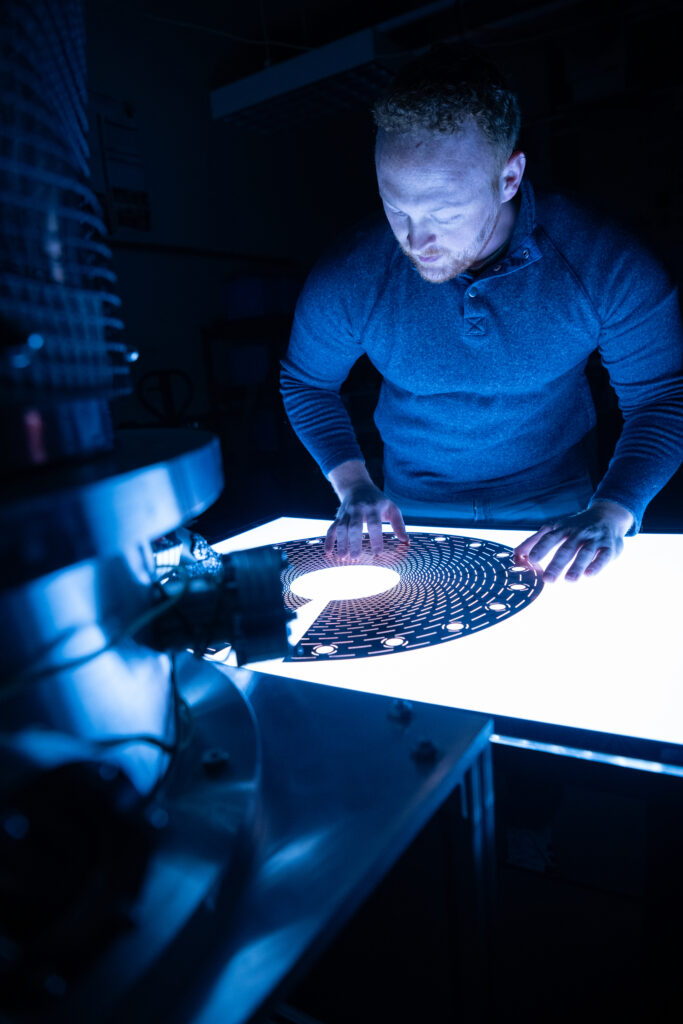 Man stands next to illuminated metal disk etched with holes.