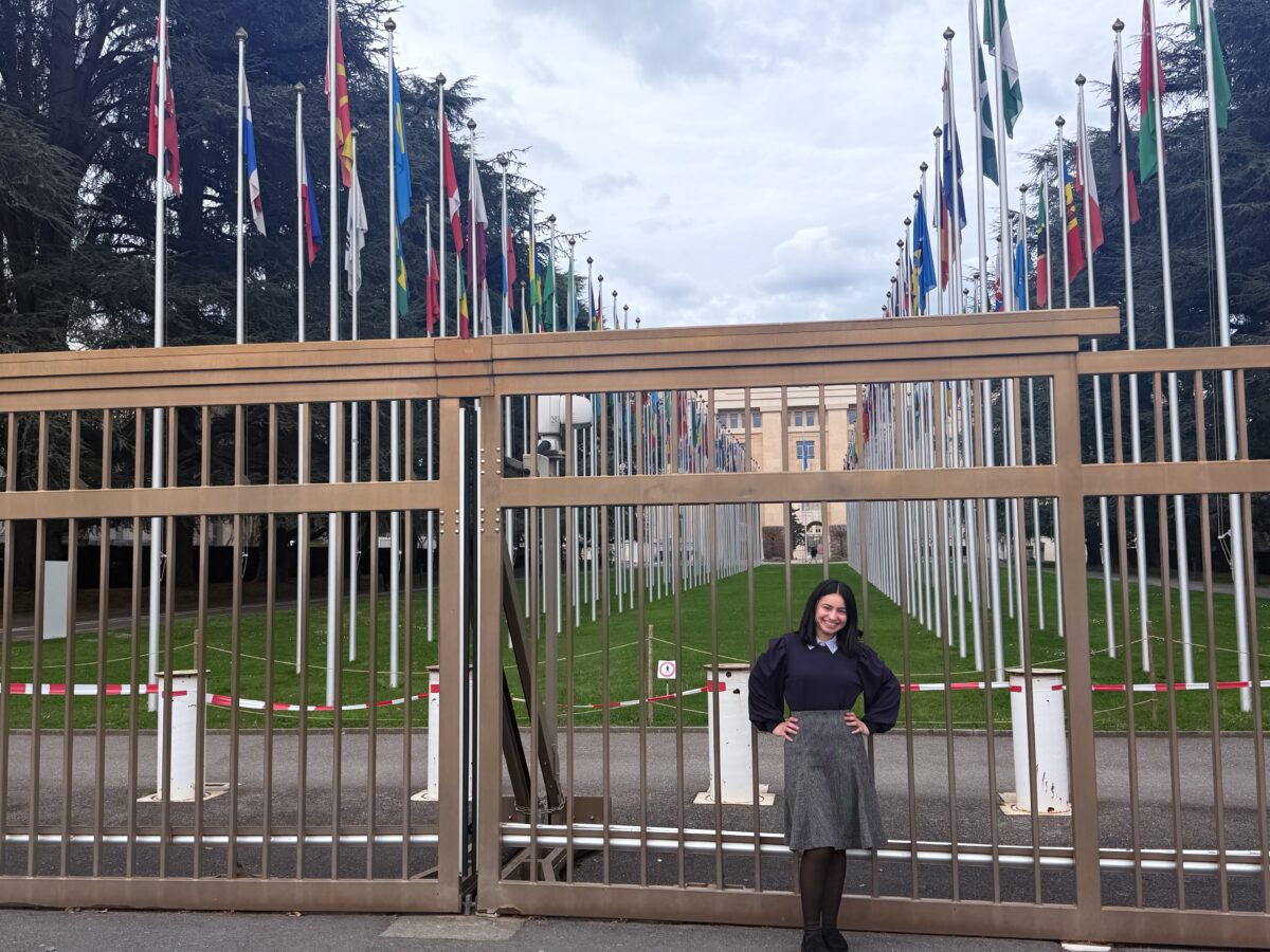 College student, Magaly Lizama Hernandez, a Bernard L. Berkowitz Memorial Award recipient, stands in front of a tall steel door that guards a government building in Switzerland with two rows of flags from around the world in front