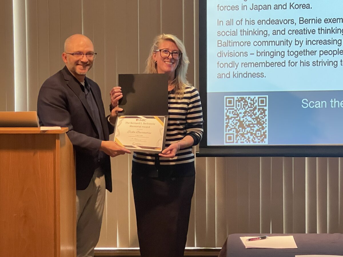 A college professor presents an award certificate to Erika Bucciantini, a doctoral student, in front of a lectern with a projection screen in the background with text about the award