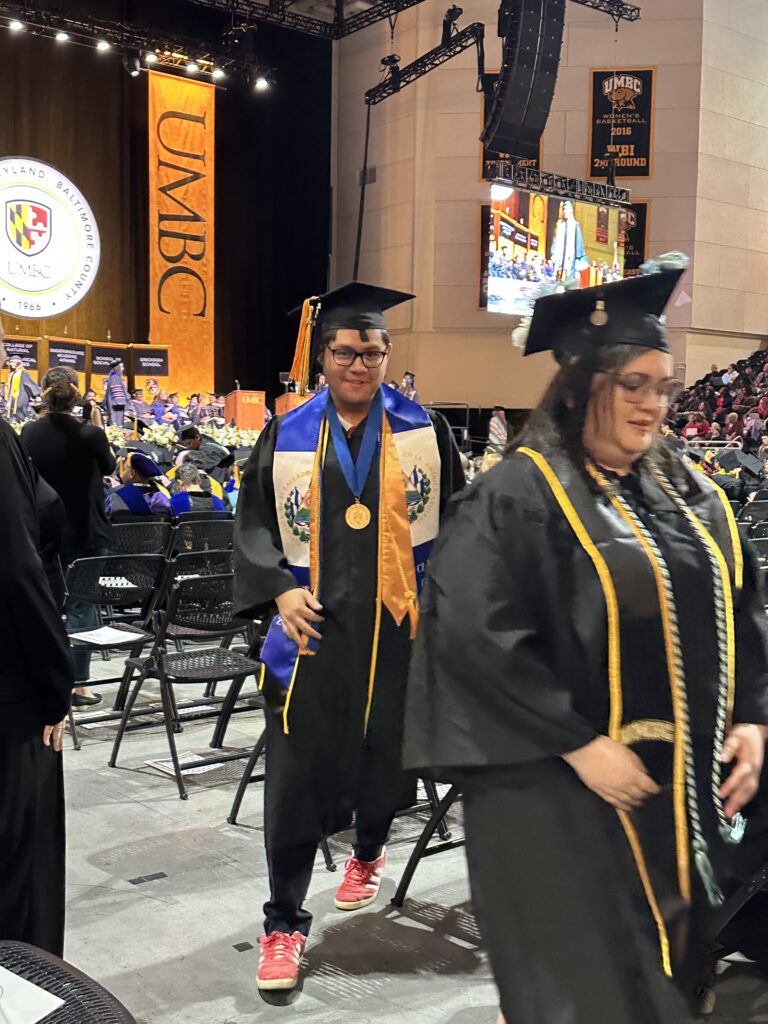 Balmory Moran, a college student, wearing his black graduation cap and gown and blue and white stole and a gold stole as he walks behind a student towards the stage