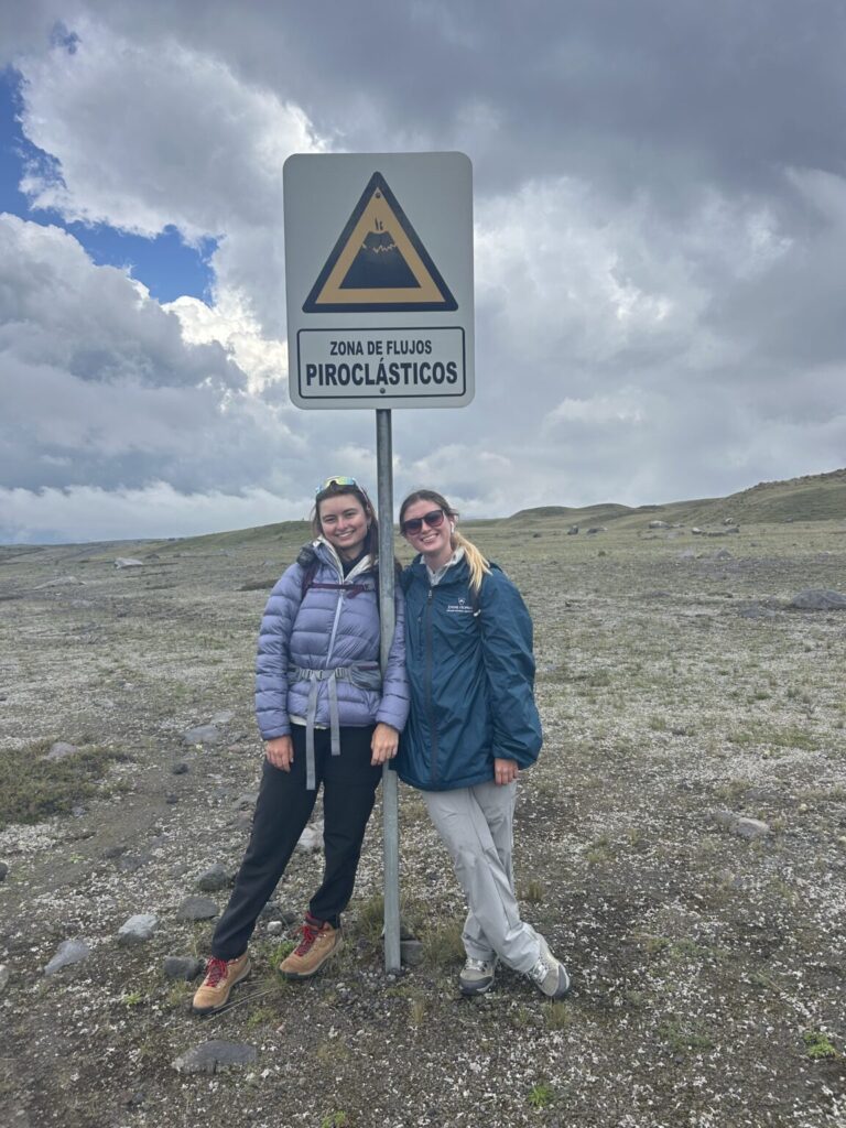 two women lean up against a sign in spanish that warns of vulcanos