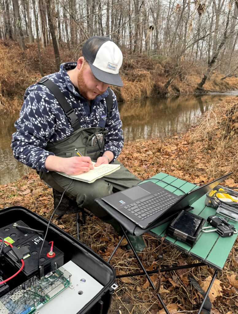 Payton Barry, wearing waders, writes in a notebook, surrounded by electronic devices and a laptop on a small foldable camping table