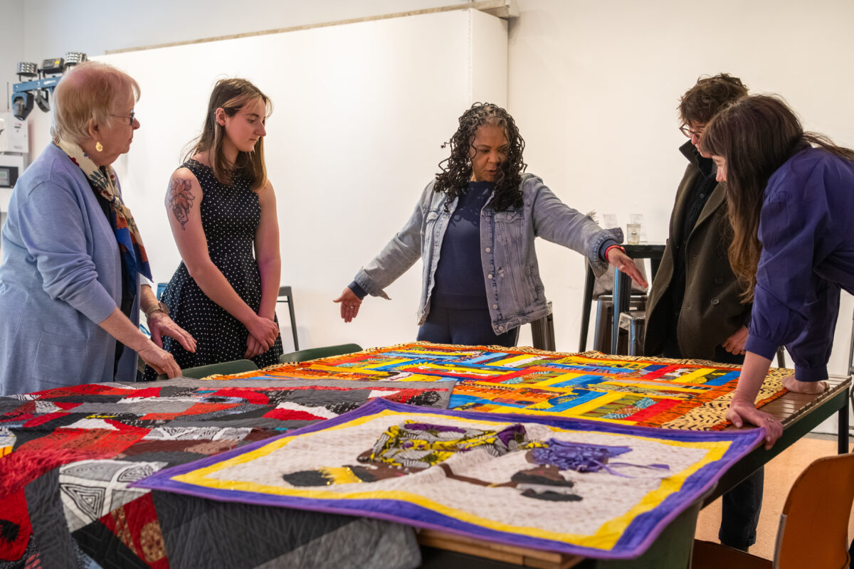 A group of five people stand around  a table inspecting multiple colorful quilts