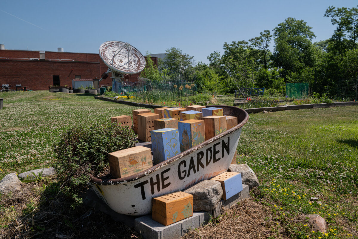 an old boat on land filled with bee hotels is decorated by text that says The Garden.