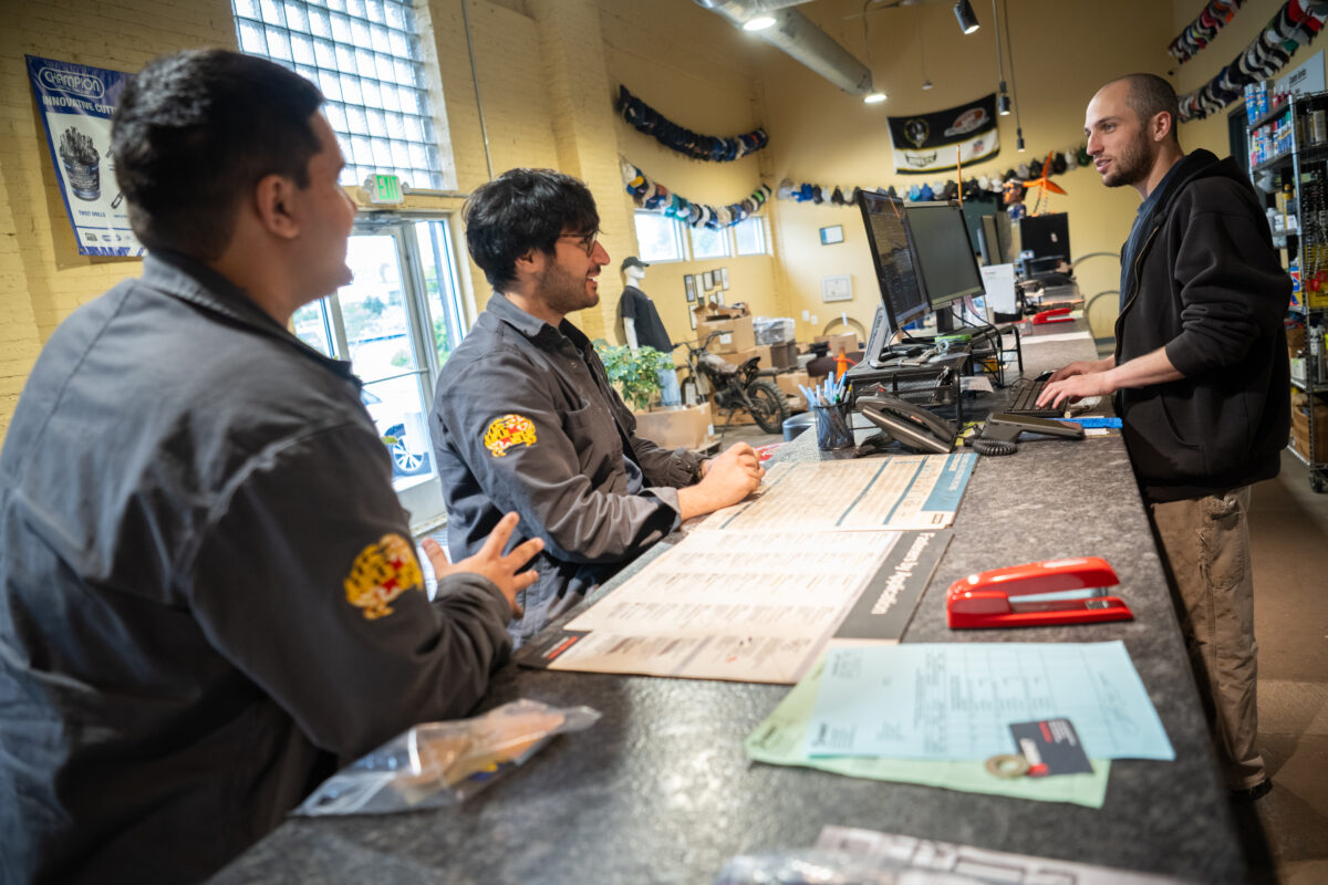 Two students in UMBC racing shirts talk to man behind the counter in a shop.